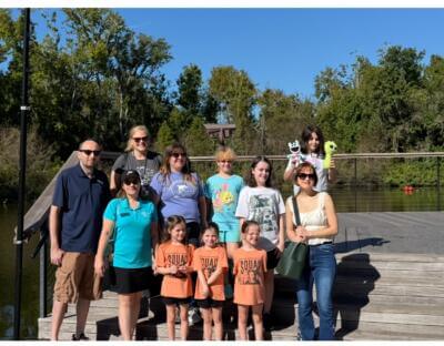 Yon gwoup moun, ki gen ladan l granmoun ak timoun, kanpe ap souri sou yon teras an bwa bò yon lak.