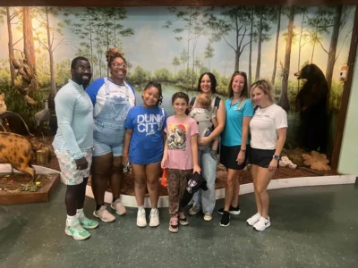 Sèt moun, ki gen ladan de timoun, kanpe anndan kay la ap souri, devan yon miray ki gen tèm forè ak figi bèt.