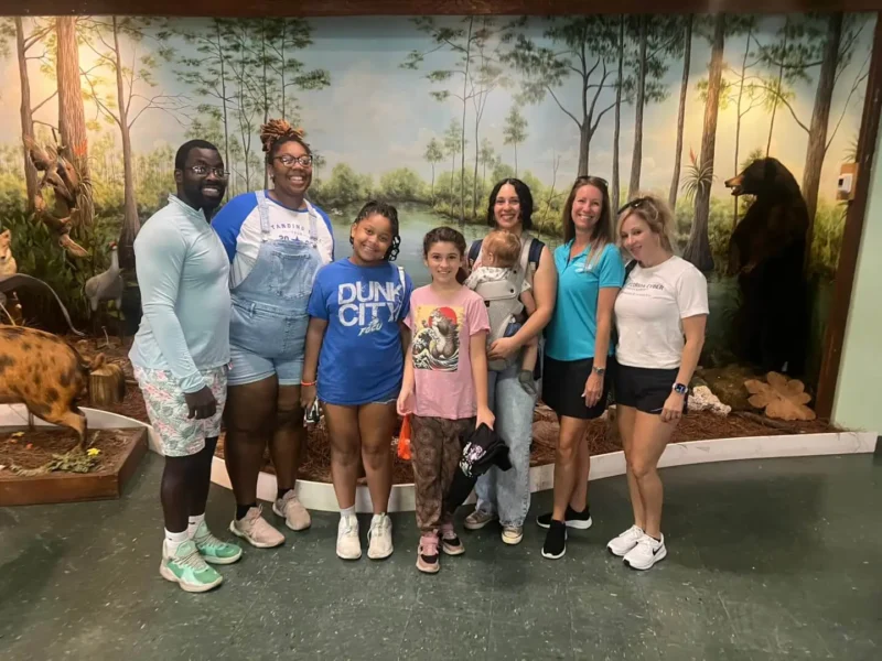 Sèt moun, ki gen ladan de timoun, kanpe anndan kay la ap souri, devan yon miray ki gen tèm forè ak figi bèt.