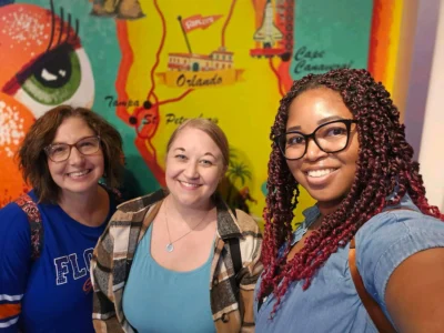 Twa fanm k ap souri devan yon miray ak yon kat jeyografik kolore ki gen moniman Florid.