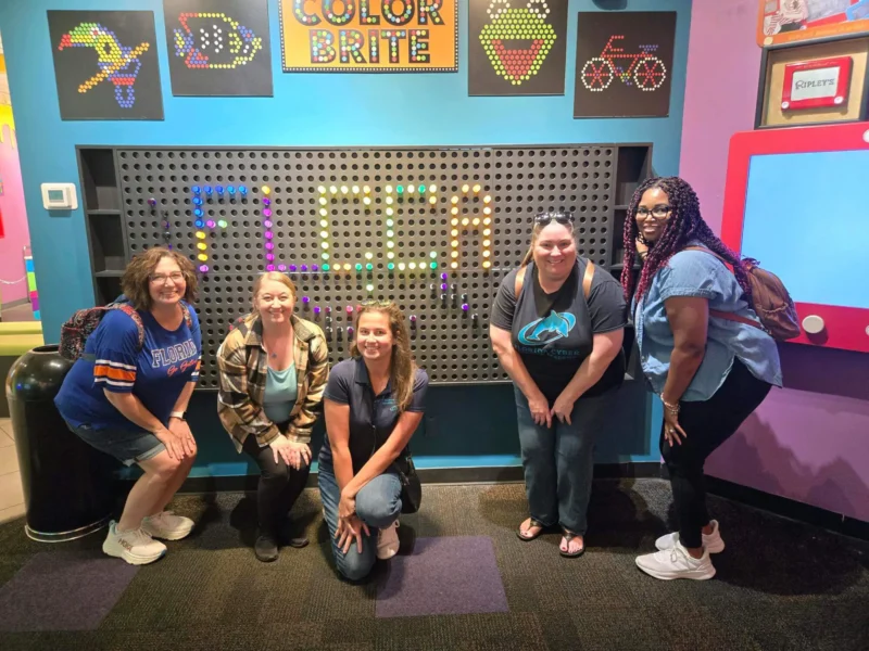 Senk moun poze ap souri devan yon ekran entèaktif kolore ak "FLCCA" ekri an limyè.